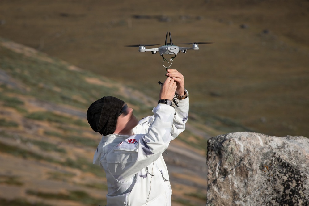 A person wearing a white jacket and black beanie hat is launching or catching a small consumer drone (appears to be a DJI Mavic Mini or similar model) with both hands raised overhead. The setting appears to be an outdoor, open terrain environment with rocky and grassy landscape in the background. The drone's propellers appear to be spinning, suggesting it is either being launched or retrieved.