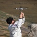 A person wearing a white jacket and black beanie hat is launching or catching a small consumer drone (appears to be a DJI Mavic Mini or similar model) with both hands raised overhead. The setting appears to be an outdoor, open terrain environment with rocky and grassy landscape in the background. The drone's propellers appear to be spinning, suggesting it is either being launched or retrieved.