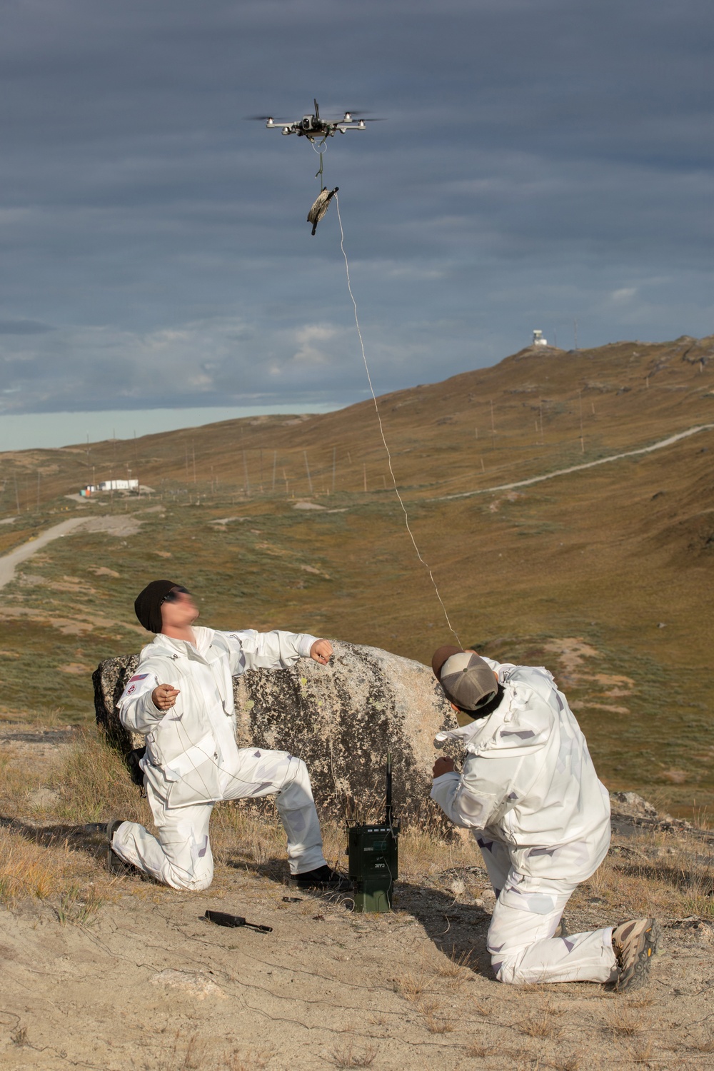 Two individuals wearing white coverall suits are kneeling behind a large boulder in an open, hilly terrain. A quadcopter drone is hovering overhead, connected by a thin line or wire to what appears to be a payload suspended beneath it. The scene suggests a drone delivery or retrieval operation, possibly in a training or field exercise context.