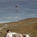Two individuals wearing white coverall suits are kneeling behind a large boulder in an open, hilly terrain. A quadcopter drone is hovering overhead, connected by a thin line or wire to what appears to be a payload suspended beneath it. The scene suggests a drone delivery or retrieval operation, possibly in a training or field exercise context.