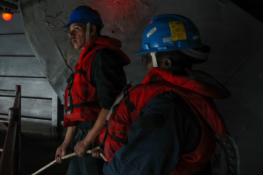 Two workers wearing blue hard hats and red life vests/personal flotation devices are positioned in what appears to be a confined or below-deck maritime environment. The person in the foreground is holding a rope or line, and both individuals appear alert and focused. The setting suggests a maritime safety drill, vessel operation, or docking procedure.