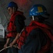 Two workers wearing blue hard hats and red life vests/personal flotation devices are positioned in what appears to be a confined or below-deck maritime environment. The person in the foreground is holding a rope or line, and both individuals appear alert and focused. The setting suggests a maritime safety drill, vessel operation, or docking procedure.