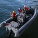 An aerial view of a gray rigid inflatable boat (RIB) being hoisted or lowered by ropes/davit lines over open water. Several personnel wearing red life vests and colored hard hats are aboard the vessel, with one individual appearing to signal with their hand. The boat displays an American flag and markings consistent with U.S. Navy equipment.