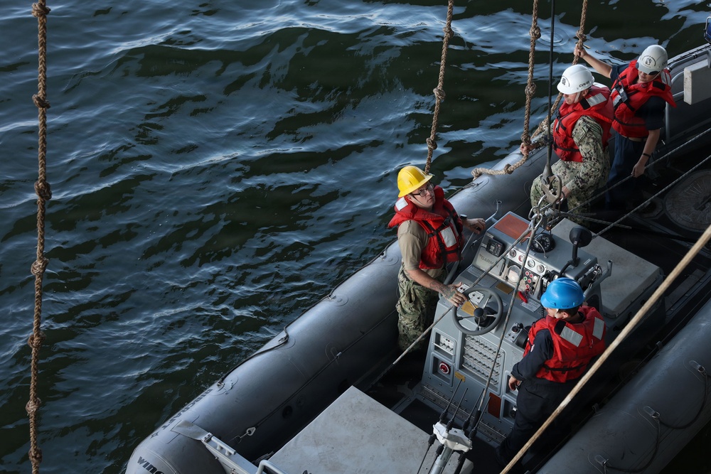 A group of four military personnel, likely U.S. Navy sailors, are operating a rigid-hull inflatable boat (RHIB) on open water. The personnel are wearing standard naval working uniforms, red personal flotation devices (life vests), and colored hard hats indicating different roles. The image is taken from an elevated angle, likely from the deck of a larger vessel, showing ropes/lines used for boat handling.