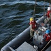 A group of four military personnel, likely U.S. Navy sailors, are operating a rigid-hull inflatable boat (RHIB) on open water. The personnel are wearing standard naval working uniforms, red personal flotation devices (life vests), and colored hard hats indicating different roles. The image is taken from an elevated angle, likely from the deck of a larger vessel, showing ropes/lines used for boat handling.