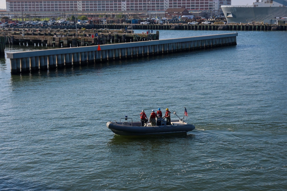 A rigid inflatable boat (RIB) with approximately four crew members wearing red life vests and hard hats is navigating a harbor waterway. An American flag is mounted on the stern of the vessel. In the background, a naval pier facility is visible with what appears to be a large warship docked at the far right.