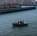A rigid inflatable boat (RIB) with approximately four crew members wearing red life vests and hard hats is navigating a harbor waterway. An American flag is mounted on the stern of the vessel. In the background, a naval pier facility is visible with what appears to be a large warship docked at the far right.
