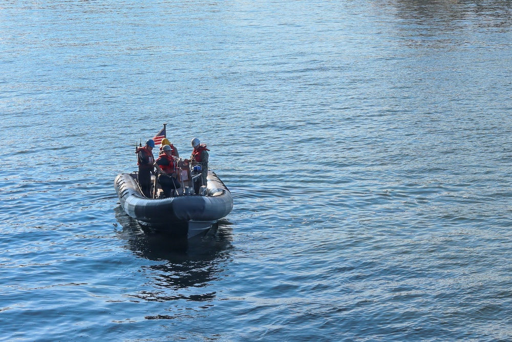A rigid inflatable boat (RIB) with approximately three personnel is operating on calm blue water. The crew members are wearing safety gear including life vests and helmets, and a small American flag is visible mounted on the vessel. The image appears to be taken from an elevated vantage point looking down at the boat.