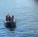 A rigid inflatable boat (RIB) with approximately three personnel is operating on calm blue water. The crew members are wearing safety gear including life vests and helmets, and a small American flag is visible mounted on the vessel. The image appears to be taken from an elevated vantage point looking down at the boat.