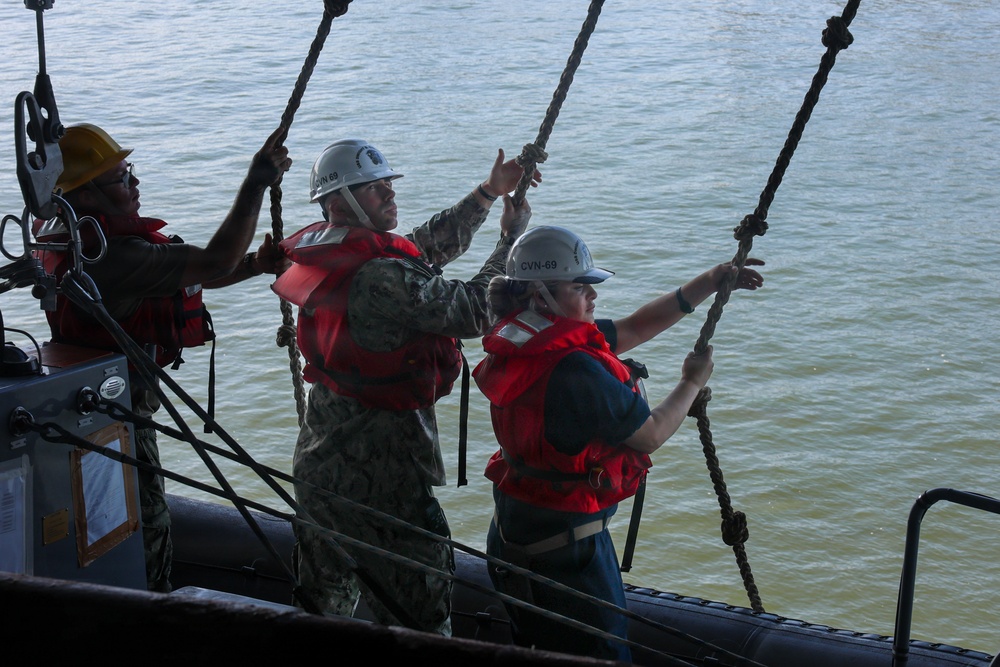 Three military personnel are working together on what appears to be a naval vessel, handling multiple ropes over water. Two individuals are wearing white hard hats marked 'CVN-69' and red life vests, while a third wears a yellow hard hat. They appear to be conducting a line-handling or rigging operation from the deck of a ship.