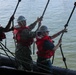 Three military personnel are working together on what appears to be a naval vessel, handling multiple ropes over water. Two individuals are wearing white hard hats marked 'CVN-69' and red life vests, while a third wears a yellow hard hat. They appear to be conducting a line-handling or rigging operation from the deck of a ship.