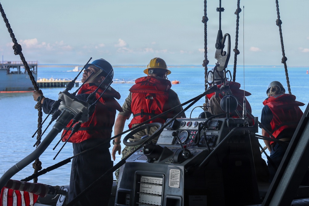 Military or coast guard personnel are operating a small tactical watercraft, likely during a training exercise or operational deployment. The crew members are wearing red life vests and safety helmets, positioned around a console equipped with a Garmin navigation system. A pier and open water are visible in the background.