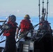 Military or coast guard personnel are operating a small tactical watercraft, likely during a training exercise or operational deployment. The crew members are wearing red life vests and safety helmets, positioned around a console equipped with a Garmin navigation system. A pier and open water are visible in the background.