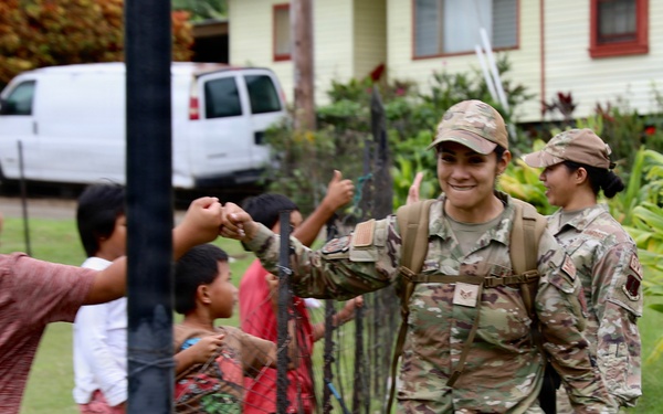 Two military personnel in OCP (Operational Camouflage Pattern) uniforms are walking along a fence line while interacting with a group of children on the other side. The lead soldier is smiling and appears to be fist-bumping or high-fiving the children through the fence. The setting appears to be a tropical or subtropical residential area based on the lush vegetation and building style visible in the background.