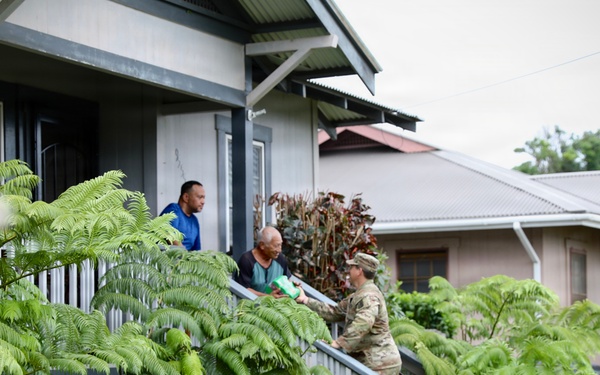 A military or National Guard personnel in camouflage uniform is interacting with two civilians on the porch of a residential home, appearing to hand over or exchange documents or materials. The setting appears to be a tropical or subtropical neighborhood, likely Hawaii based on the lush tree ferns and architectural style. Additional uniformed personnel are visible in the foreground, suggesting an organized outreach or welfare check operation.