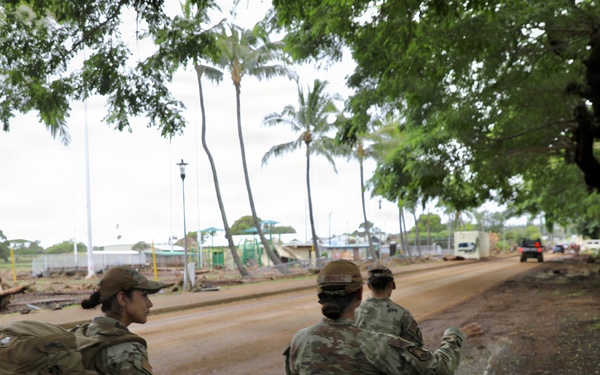 Three military personnel in camouflage uniforms are walking along a mud-covered road in what appears to be a disaster response scenario. The area shows significant debris and mud deposits consistent with flooding or a natural disaster, with palm trees and damaged structures visible in the background. One soldier is gesturing forward, suggesting they are conducting an assessment or search operation.