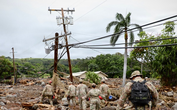 A group of military personnel in camouflage uniforms traverse a heavily debris-strewn landscape following what appears to be a flash flood or mudslide disaster. The scene shows extensive destruction including displaced boulders, uprooted trees, scattered debris, and downed power lines with leaning utility poles. The tropical vegetation in the background and red-brown mud suggest a Hawaiian or similar Pacific island location.