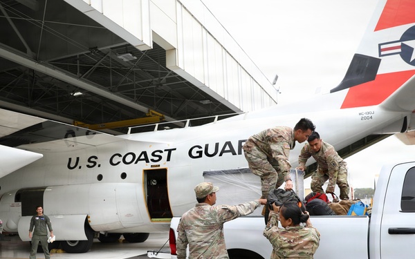Military personnel in camouflage uniforms are loading or unloading equipment and bags from a white pickup truck onto or from a U.S. Coast Guard aircraft on a tarmac. Several service members in flight suits are observing nearby. The scene appears to be a logistics or deployment operation at a Coast Guard air station.