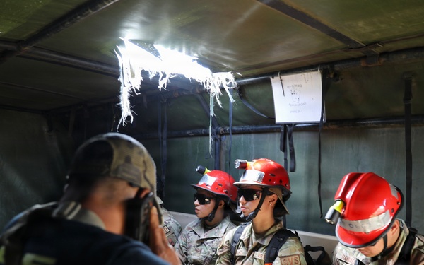 A group of military personnel wearing camouflage uniforms and red safety helmets with face shields are seated inside what appears to be a covered military vehicle or tent structure. At least one civilian in a dark shirt and baseball cap is present, appearing to brief or communicate with the uniformed personnel. A patch reading 'SVS' is visible on multiple uniforms, and one name tape reads 'BANIABA'.
