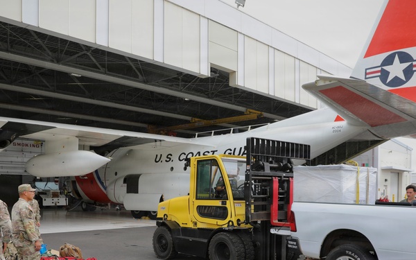 Military personnel and ground crew are loading cargo onto or near a U.S. Coast Guard aircraft at what appears to be an air station hangar. A yellow forklift is transferring a wrapped pallet from a white pickup truck, while soldiers in camouflage uniforms stand nearby with red duffel bags and equipment on the tarmac. The scene suggests a logistics or deployment operation involving both Army personnel and Coast Guard assets.