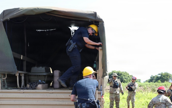 The image shows a joint military and civilian emergency response exercise or operation in an outdoor field setting. Personnel in military camouflage uniforms and civilian responders wearing yellow and red hard hats are gathered around a tan military cargo truck. Two individuals in dark uniforms with hard hats are climbing into or inspecting the rear of the vehicle while soldiers observe.