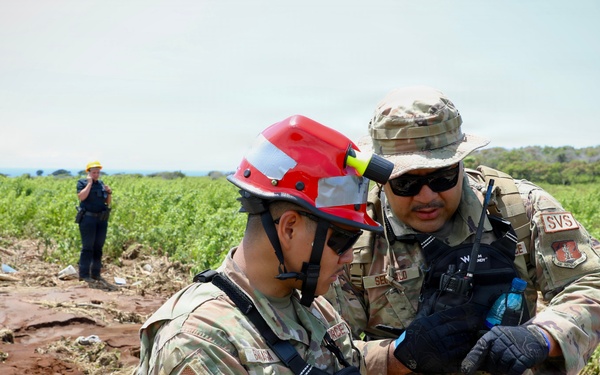 Two military personnel in camouflage uniforms are examining a mobile device at what appears to be a disaster response or search-and-rescue site with disturbed red soil and debris. One individual is wearing a red rescue helmet with a mounted flashlight, while the other wears a boonie hat and sunglasses with 'SVS' patch visible. A third person in the background, wearing a yellow hard hat, observes from a distance.
