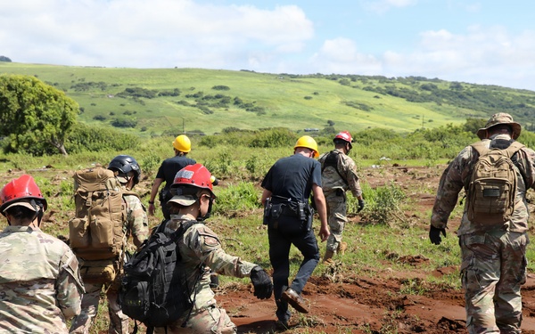 A group of military personnel and civilian emergency responders are traversing a muddy, cracked terrain in what appears to be a post-disaster search and rescue operation. The team is moving away from the camera across red clay mud toward a green hillside, suggesting a flood or landslide response scenario. Personnel are wearing a mix of military camouflage uniforms and civilian emergency gear with colored hard hats.