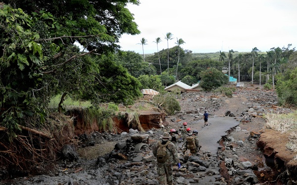 The image shows a disaster response scene where a group of personnel in military-style uniforms and red helmets are walking through a heavily flood-damaged area. A road or pathway has been severely eroded, leaving exposed rock, mud, and debris on both sides. Lush tropical vegetation and palm trees are visible in the background, suggesting a tropical or subtropical location.