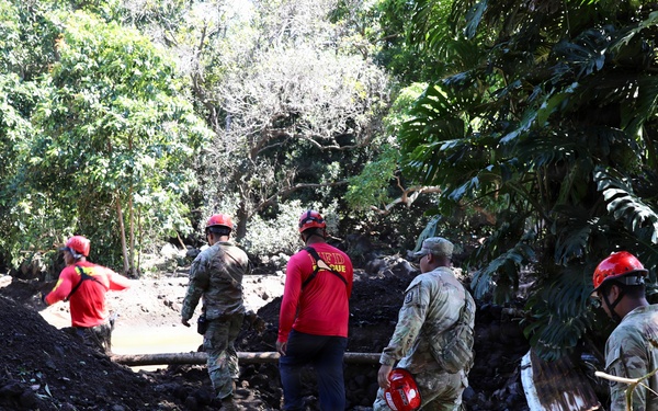 A group of rescue and military personnel are working at what appears to be a disaster site, likely following a landslide or flood event. Workers in camouflage uniforms and red helmets are navigating through displaced earth, debris, and fallen branches. The scene suggests an active search and rescue or debris clearance operation in a tropical or subtropical environment.