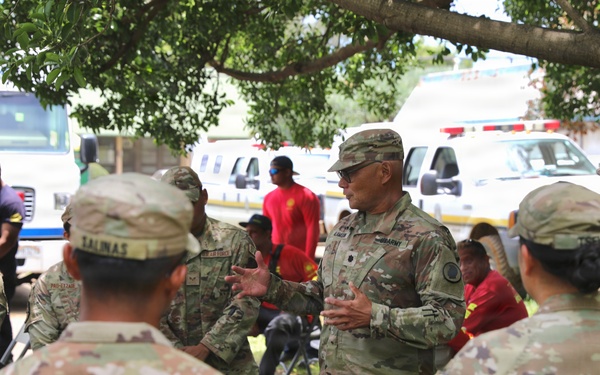 A group of U.S. military personnel in OCP (Operational Camouflage Pattern) uniforms are gathered in an outdoor briefing or debriefing session under a tree. A central figure, identifiable as a senior officer, appears to be addressing the group. Emergency or support vehicles are visible in the background, suggesting a disaster response or field operation context.