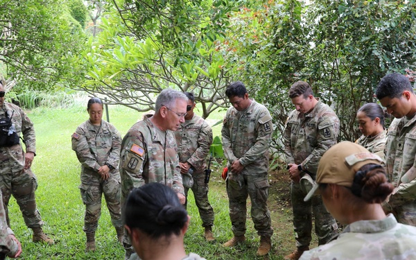 A group of U.S. Army soldiers in OCP (Operational Camouflage Pattern) uniforms are gathered in a circle outdoors in a green, vegetated area. The soldiers appear to be bowing their heads in what appears to be a prayer or moment of reflection. A senior officer, identifiable by rank insignia, appears to be leading the group.