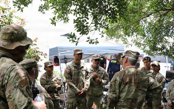 A group of U.S. Army soldiers in OCP (Operational Camouflage Pattern) uniforms are gathered in an outdoor briefing or debriefing circle under tree cover. The setting appears to be a field operation or disaster response staging area, indicated by pop-up canopy tents and equipment visible in the background. One soldier appears to be addressing the group in a leadership capacity.