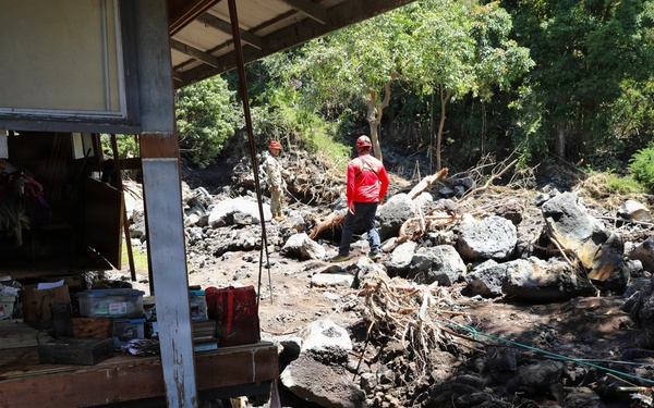 The image depicts the aftermath of a landslide or flash flood event that has caused significant destruction to a residential structure. Large boulders, debris, mud, uprooted trees, and displaced household items are scattered across the affected area. Two individuals wearing red clothing and caps appear to be surveying or assessing the damage.