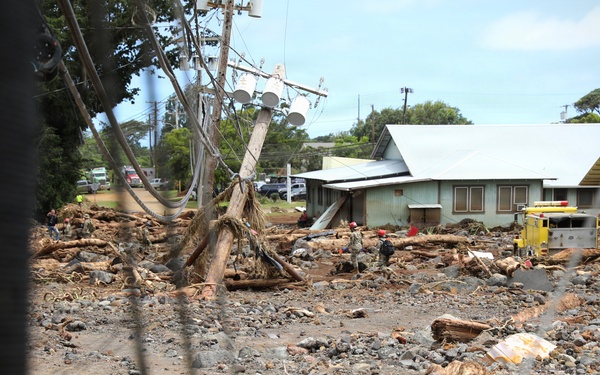 The image depicts a residential area severely impacted by what appears to be a flash flood or debris flow event. Large amounts of rocks, mud, uprooted trees, and debris are scattered across what was likely a road or open area between structures. Emergency responders, including at least one fire/rescue vehicle, are visible in the background conducting operations.