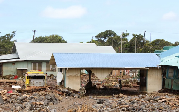 The image depicts severe flood and debris flow damage to a residential area, with large rocks, logs, mud, and debris covering the ground surrounding several structures. A yellow emergency vehicle (likely a fire truck) is visible among the damaged buildings. The scene suggests a recent flash flood or debris flow event that has inundated the neighborhood.