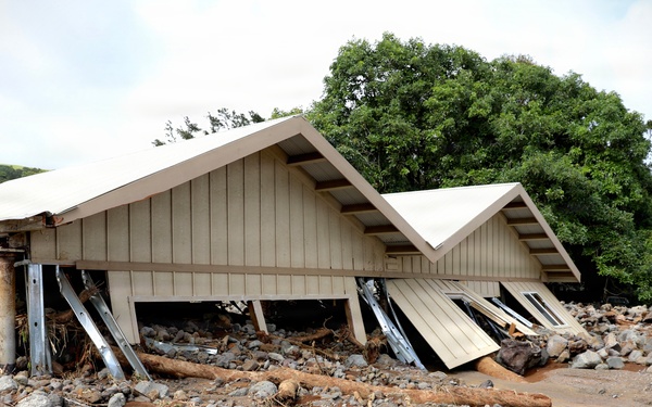 A building has been severely damaged by what appears to be a flash flood or debris flow event. The structure's walls have collapsed or been pushed inward while the roof remains partially intact, supported by metal posts. The surrounding area is covered in a thick layer of rocks, mud, debris, and uprooted logs consistent with a high-energy flood or landslide event.