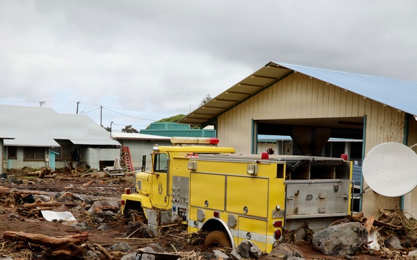 The image shows significant flood and debris damage to a small community, with a yellow emergency/fire truck partially buried in mud, rocks, and debris. Large logs, boulders, and sediment-laden floodwater remnants cover the ground around residential and utility buildings. A satellite dish is visible on the right side of a damaged structure.