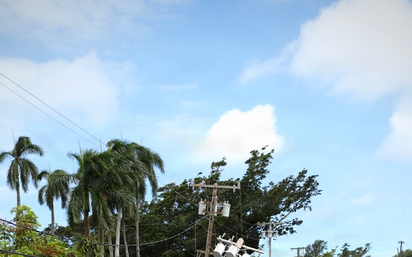 The image shows severe flood or storm damage to what appears to be a tropical residential area, likely in Hawaii based on the palm trees and volcanic rock. A utility pole with transformers has been knocked over at an angle, with downed power lines draped across a debris-covered landscape. The ground is blanketed with rocks, logs, mud, and scattered debris, indicating a recent flash flood or major storm event.