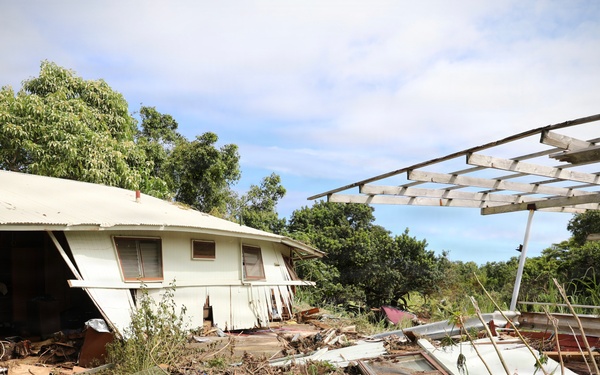 The image shows a severely damaged residential property, likely following a flood or storm event. A white single-story house has sustained significant structural damage, with debris, mud, and displaced materials scattered across the surrounding ground. A metal carport or pergola frame on the right side has also been damaged, with its panels stripped away.