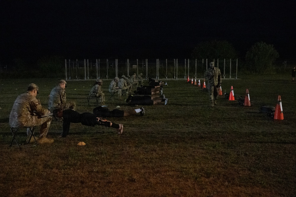 A nighttime military physical fitness test is being conducted on a grass field. Personnel in civilian workout attire are performing push-ups while uniformed soldiers seated on folding chairs observe and record scores. Orange traffic cones mark lanes or boundaries along the right side of the field.