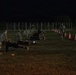 A nighttime military physical fitness test is being conducted on a grass field. Personnel in civilian workout attire are performing push-ups while uniformed soldiers seated on folding chairs observe and record scores. Orange traffic cones mark lanes or boundaries along the right side of the field.