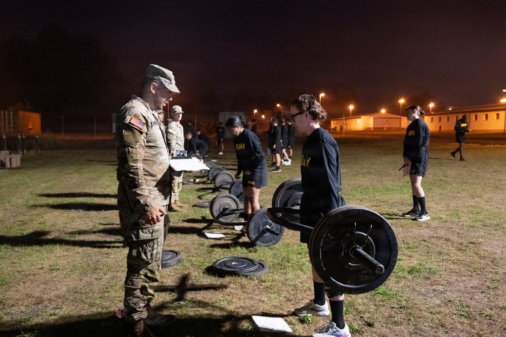 A nighttime U.S. Army physical fitness training session taking place on an outdoor field at a military installation. Soldiers in PT (physical training) black uniforms are performing deadlift exercises with barbells while a uniformed cadre member in camouflage holds a clipboard, apparently grading or supervising the event. The scene appears to be a formal fitness assessment, likely the Army Combat Fitness Test (ACFT).