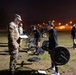 A nighttime U.S. Army physical fitness training session taking place on an outdoor field at a military installation. Soldiers in PT (physical training) black uniforms are performing deadlift exercises with barbells while a uniformed cadre member in camouflage holds a clipboard, apparently grading or supervising the event. The scene appears to be a formal fitness assessment, likely the Army Combat Fitness Test (ACFT).