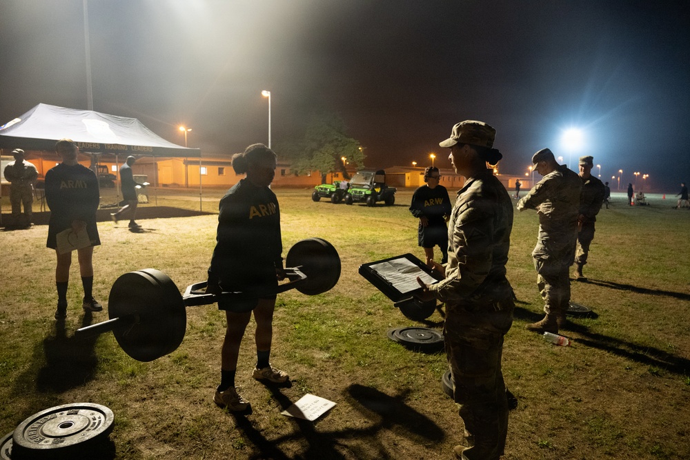 A nighttime U.S. Army physical fitness assessment is taking place on an outdoor field, illuminated by stadium-style floodlights. A soldier in Army PT (physical training) uniform is performing a deadlift with a loaded barbell while a uniformed evaluator holding a clipboard observes and records results. Additional soldiers and personnel are visible in the background near a white tent and utility vehicles.