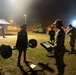 A nighttime U.S. Army physical fitness assessment is taking place on an outdoor field, illuminated by stadium-style floodlights. A soldier in Army PT (physical training) uniform is performing a deadlift with a loaded barbell while a uniformed evaluator holding a clipboard observes and records results. Additional soldiers and personnel are visible in the background near a white tent and utility vehicles.