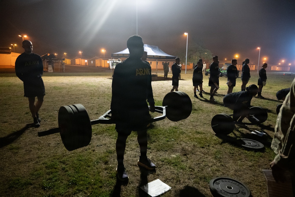 A group of U.S. Army soldiers are conducting an outdoor physical training session at night under artificial lighting. The central figure is performing or preparing to perform a deadlift using a hex/trap bar loaded with weight plates. Multiple soldiers are visible in the background engaged in various exercises on a grassy field near what appears to be a military installation.
