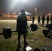 A group of U.S. Army soldiers are conducting an outdoor physical training session at night under artificial lighting. The central figure is performing or preparing to perform a deadlift using a hex/trap bar loaded with weight plates. Multiple soldiers are visible in the background engaged in various exercises on a grassy field near what appears to be a military installation.