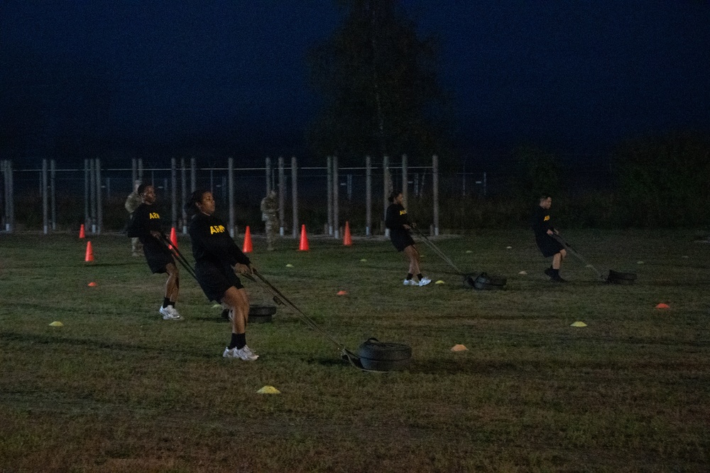 A group of U.S. Army soldiers in black PT (physical training) uniforms are conducting an early morning or evening outdoor fitness exercise on a grass field. They appear to be performing sled drag or resistance band pulling exercises, with weighted sleds visible on the ground. The training area is marked with orange traffic cones and small yellow and orange field markers.