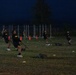A group of U.S. Army soldiers in black PT (physical training) uniforms are conducting an early morning or evening outdoor fitness exercise on a grass field. They appear to be performing sled drag or resistance band pulling exercises, with weighted sleds visible on the ground. The training area is marked with orange traffic cones and small yellow and orange field markers.