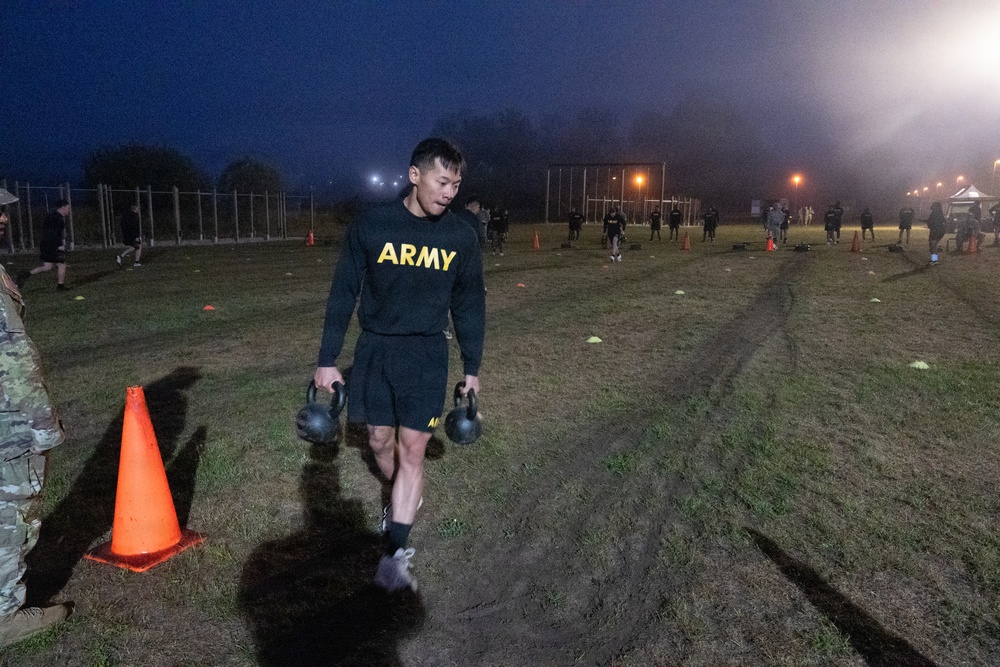 A U.S. Army soldier is performing a kettlebell farmer's carry exercise on an outdoor field during early morning or nighttime hours. The event appears to be a physical fitness training session or assessment, with multiple other soldiers visible in the background performing similar activities. Orange traffic cones mark the course lanes on a grass and dirt field illuminated by artificial lighting.